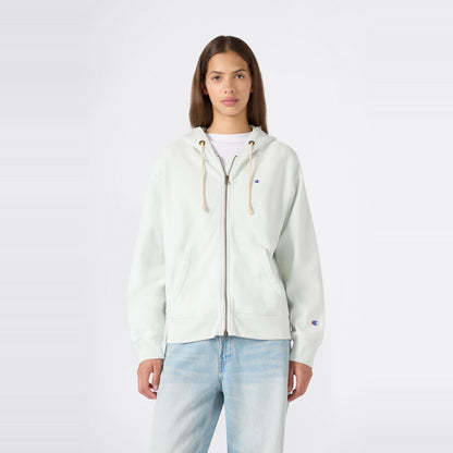 A person stands facing forward, wearing a Champion Women’s Knitted Hooded Full Zip Sweatshirt in a light color over a white shirt and paired with light blue jeans, against a plain white background.