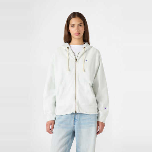 A person stands facing forward, wearing a Champion Women’s Knitted Hooded Full Zip Sweatshirt in a light color over a white shirt and paired with light blue jeans, against a plain white background.