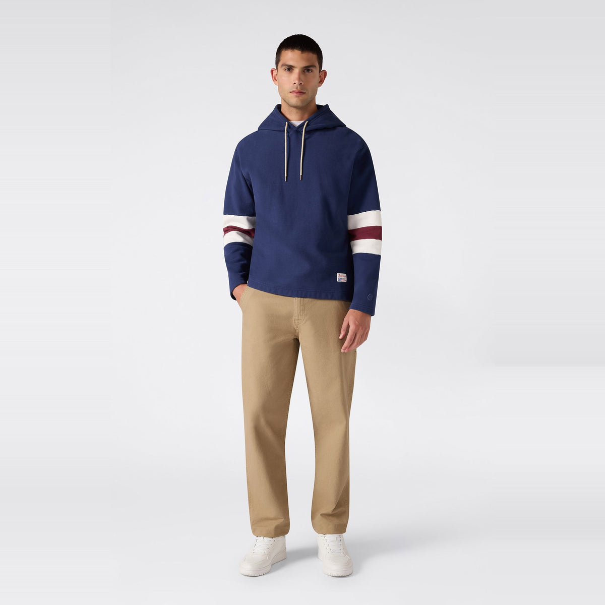 A man stands facing forward, wearing a Champion Men’s Knitted Hooded Sweatshirt in navy with white and maroon stripes on the sleeves, khaki pants, and white sneakers against a plain light background.