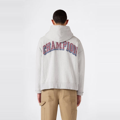 A person wearing a light gray Champion Men’s Knitted Hooded Sweatshirt (College Graphic) featuring CHAMPION in large letters on the back, standing facing away. This men’s college-inspired hoodie is crafted from soft knit fabric and is paired with tan cargo pants against a plain white background.