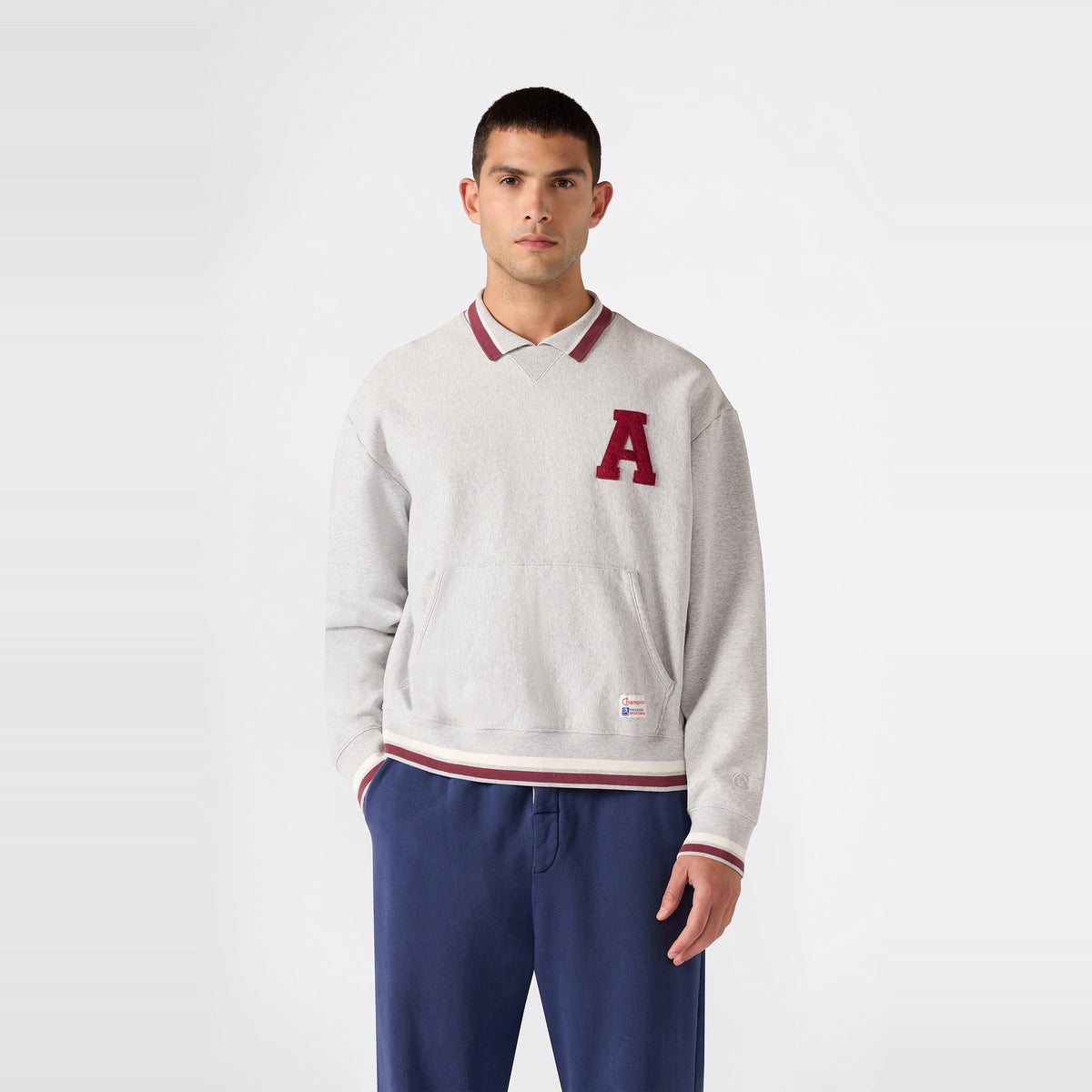 A man stands against a plain background, wearing a light gray Champion Men’s Knitted Polo Neck Sweatshirt featuring a red A on the left chest, layered over a collared shirt, and paired with blue pants.