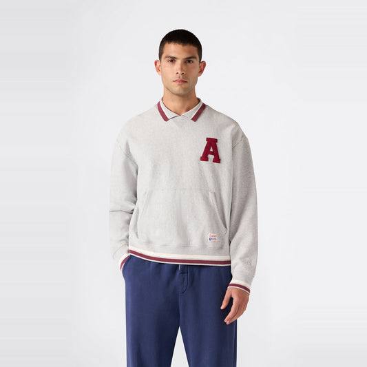 A man stands against a plain background, wearing a light gray Champion Men’s Knitted Polo Neck Sweatshirt featuring a red A on the left chest, layered over a collared shirt, and paired with blue pants.