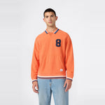 A man is wearing a Champion Men’s Knitted Polo Neck Sweatshirt in orange, featuring a blue number 8 on the chest, paired with light blue jeans, standing against a plain white background.