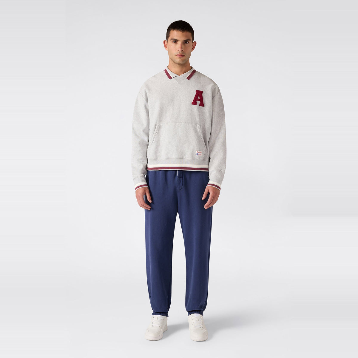 A person stands facing forward, wearing a light gray Reverse Weave fleece sweatshirt with a red A patch, paired with Champion Men’s Knitted Elastic Cuff Pants and blue and white sneakers, set against a plain light gray background.