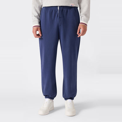 A person is wearing Champion Men’s Knitted Elastic Cuff Pants in blue, styled with white sneakers and a light gray Reverse Weave fleece sweatshirt, standing against a plain light background. Only the lower torso and legs are visible.