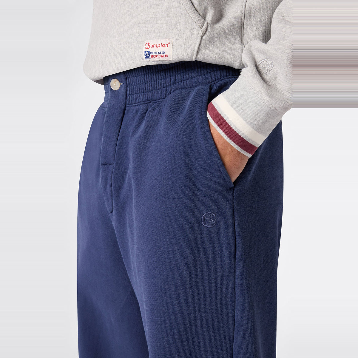A person wearing a gray Champion sweatshirt made from soft Reverse Weave fleece with striped cuffs and blue Champion Men’s Knitted Elastic Cuff Pants, one hand in the pocket, shown from the waist down against a plain background.