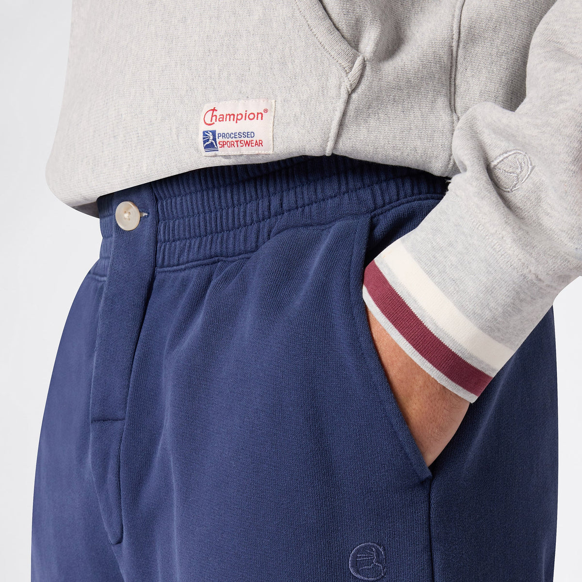 Close-up of a person wearing a grey Champion sweatshirt crafted from Reverse Weave fleece, paired with Champion Men’s Knitted Elastic Cuff Pants in navy blue, with one hand in the pocket. The Champion logo is visible on the sweatshirt above the waistband.