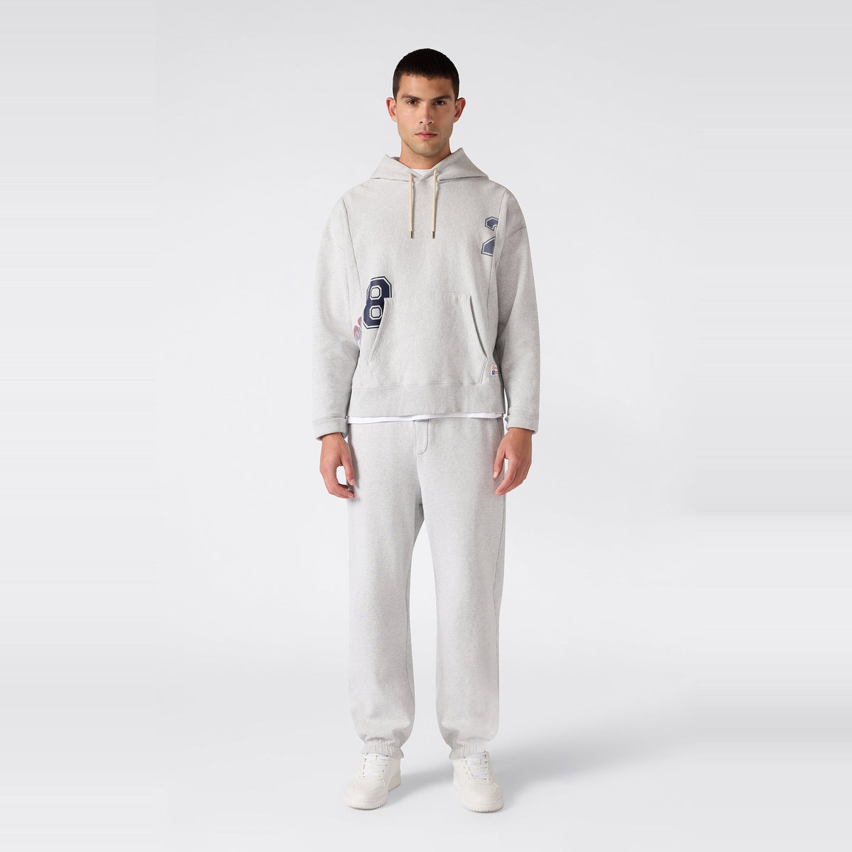 A man stands facing forward against a plain light gray background, wearing a heather gray heavyweight fleece hooded sweatshirt with navy blue “8” and “2” graphics, paired with Champion Men’s Knitted Elastic Cuff Pants in classic Reverse Weave fabric and white sneakers.