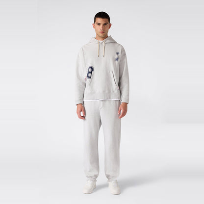 A man stands facing forward against a plain light gray background, wearing a heather gray heavyweight fleece hooded sweatshirt with navy blue “8” and “2” graphics, paired with Champion Men’s Knitted Elastic Cuff Pants in classic Reverse Weave fabric and white sneakers.