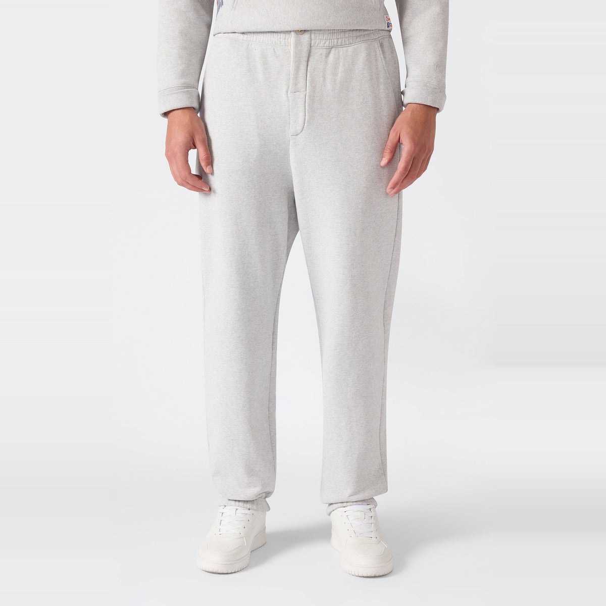 A person is shown from the waist down wearing Champion Men’s Knitted Elastic Cuff Pants in light gray, paired with white sneakers, standing against a plain white background.