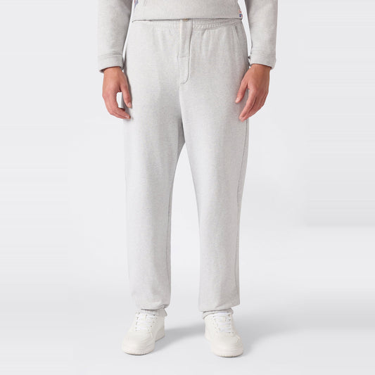 A person is shown from the waist down wearing Champion Men’s Knitted Elastic Cuff Pants in light gray, paired with white sneakers, standing against a plain white background.