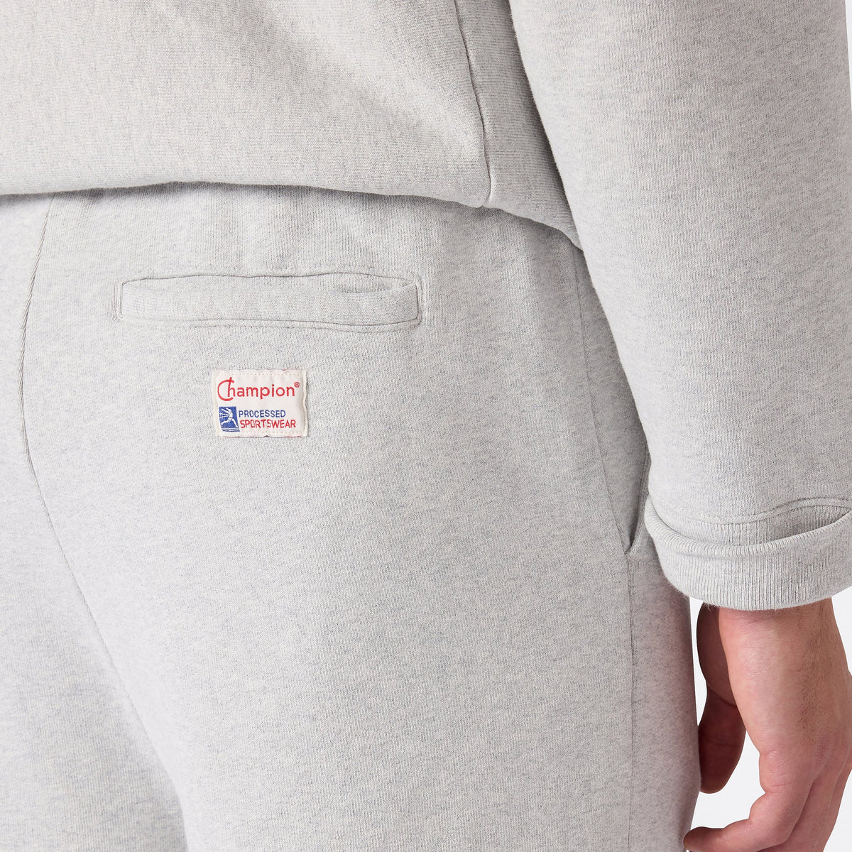Close-up of a person wearing Champion Men’s Knitted Elastic Cuff Pants in light gray, paired with a matching sweatshirt. Crafted from heavyweight fleece for superior warmth, the pants feature a rectangular Champion Processed Sportswear logo patch stitched just below the horizontal pocket flap on the back.