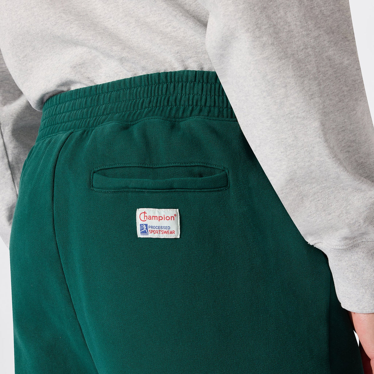 A person is wearing green Champion Men’s Knitted Elastic Cuff Pants featuring a back pocket and a white Champion logo patch, paired with a light grey long-sleeve shirt.