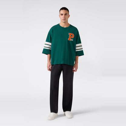 A man stands against a plain white background, wearing the Champion Men’s Knitted Crewneck T-Shirt—a relaxed fit green shirt featuring white stripes on the sleeves and a large orange letter P on the chest—paired with black pants and white sneakers.