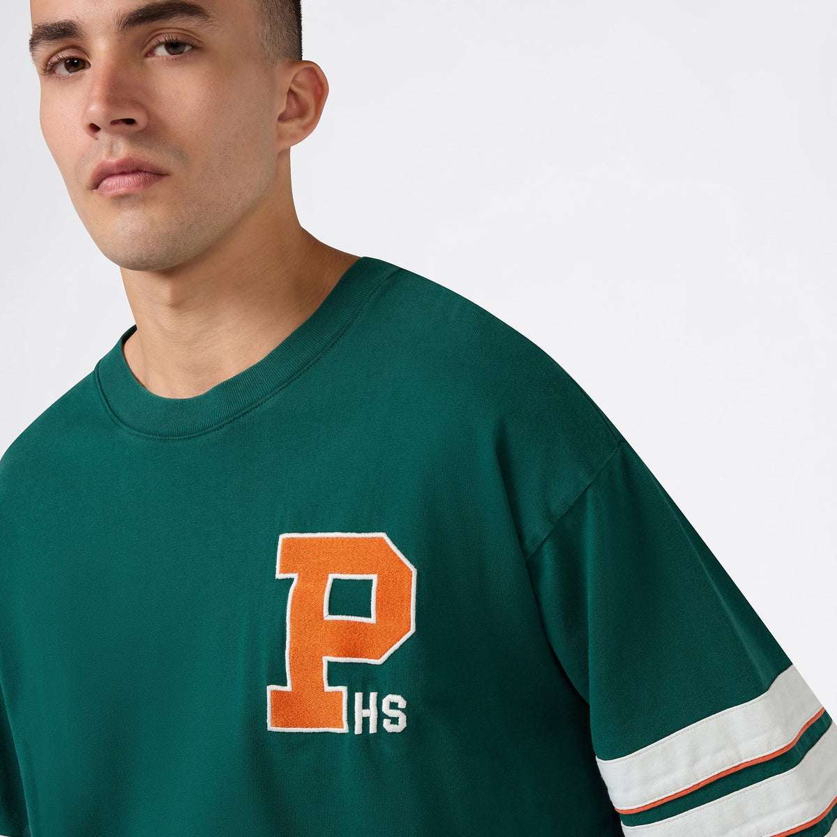 A person wearing a green Champion Men’s Knitted Crewneck T-Shirt is looking at the camera against a plain background. This crewneck features a relaxed fit for everyday comfort, with white sleeve stripes and an orange letter P above HS on the front.