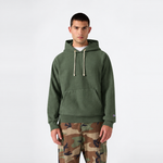 A man stands facing forward against a plain white background, wearing a green Champion Men’s Relaxed Fit Hooded Sweatshirt with drawstrings and camouflage pants.