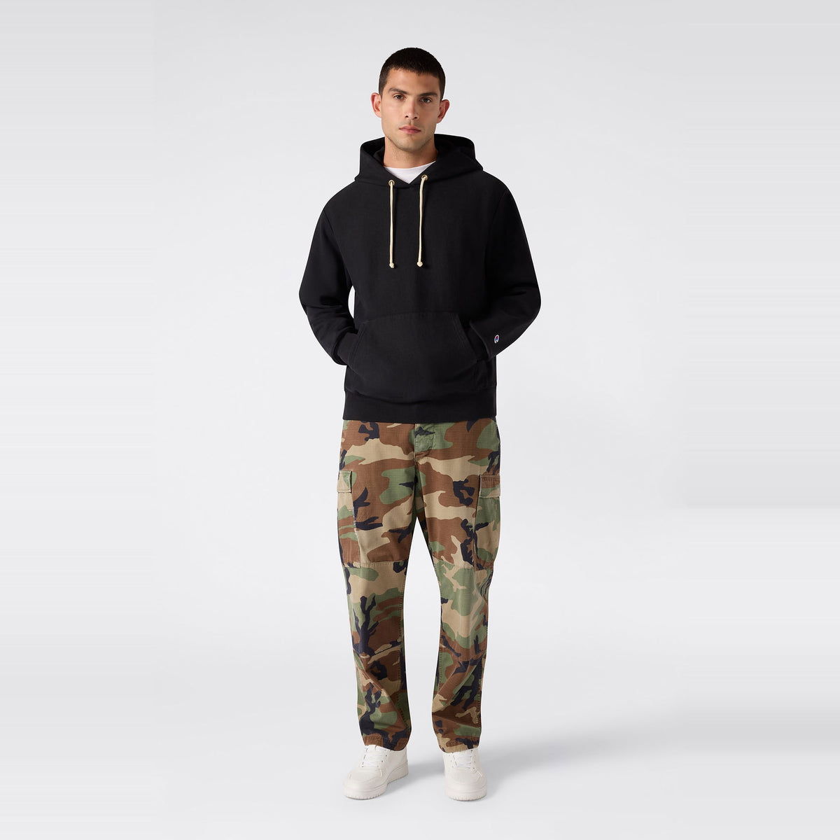 A person stands facing forward against a plain light gray background, wearing a black Champion Men’s Relaxed Fit Hooded Sweatshirt made from heavyweight fleece, paired with camouflage pants and white sneakers.