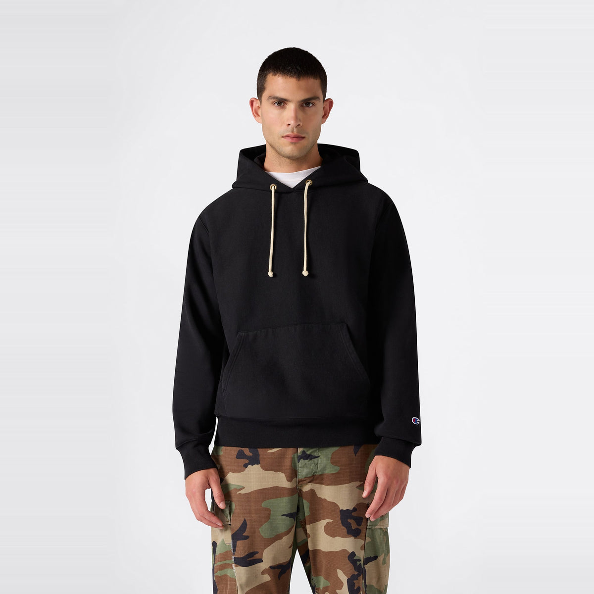 A person wearing the Champion Men’s Relaxed Fit Hooded Sweatshirt in black with beige drawstrings and camouflage pants stands against a plain white background.