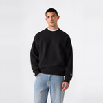 A man stands against a plain white background, wearing the Champion Men’s Knitted Crewneck Sweatshirt in black over a white shirt and light blue jeans, with one hand in his pocket. This classic sweatshirt showcases the timeless style that Champion is known for.