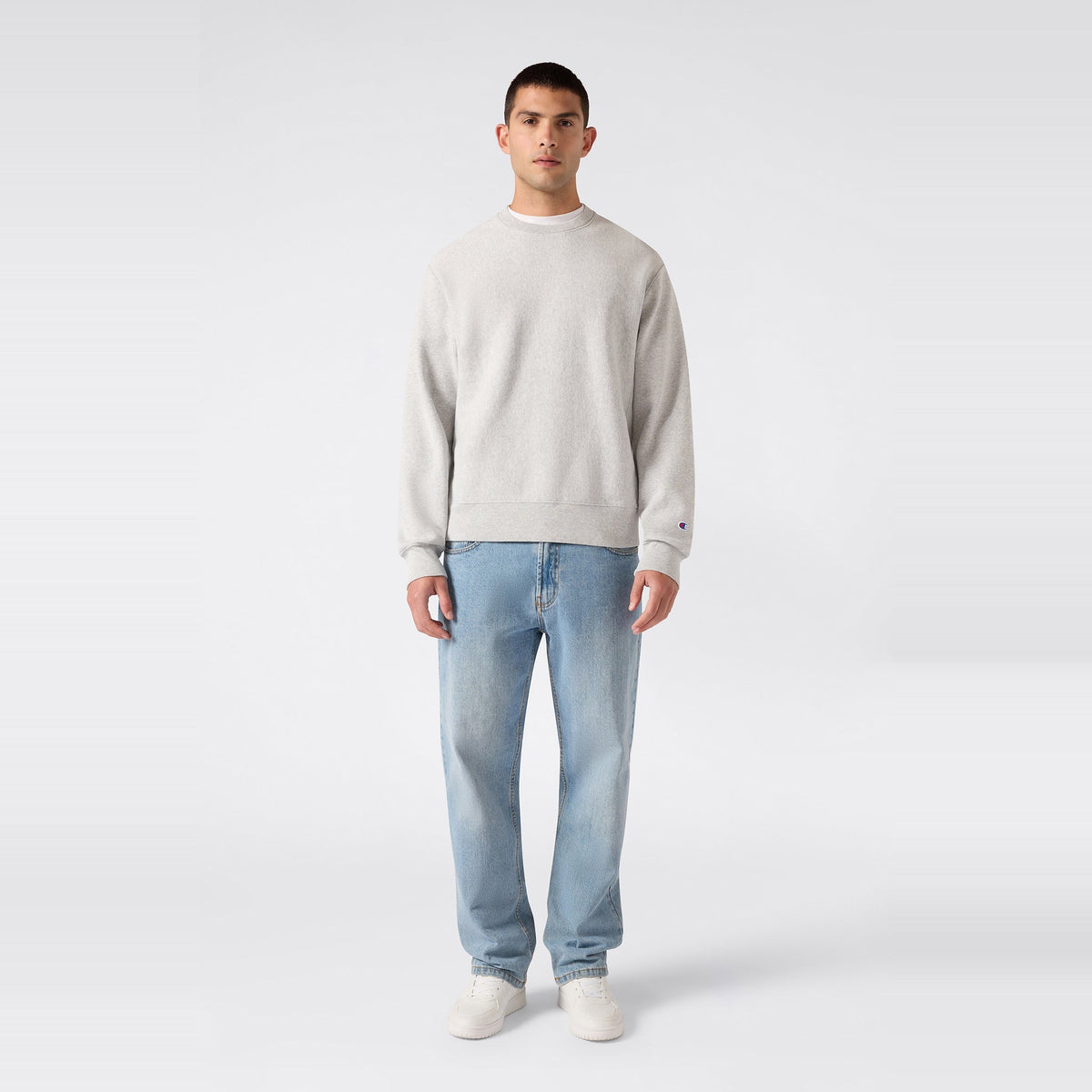 A person stands facing forward, wearing a light gray Champion Men’s Relaxed Fit Crewneck Sweatshirt, light blue jeans, and white sneakers, against a plain white background.