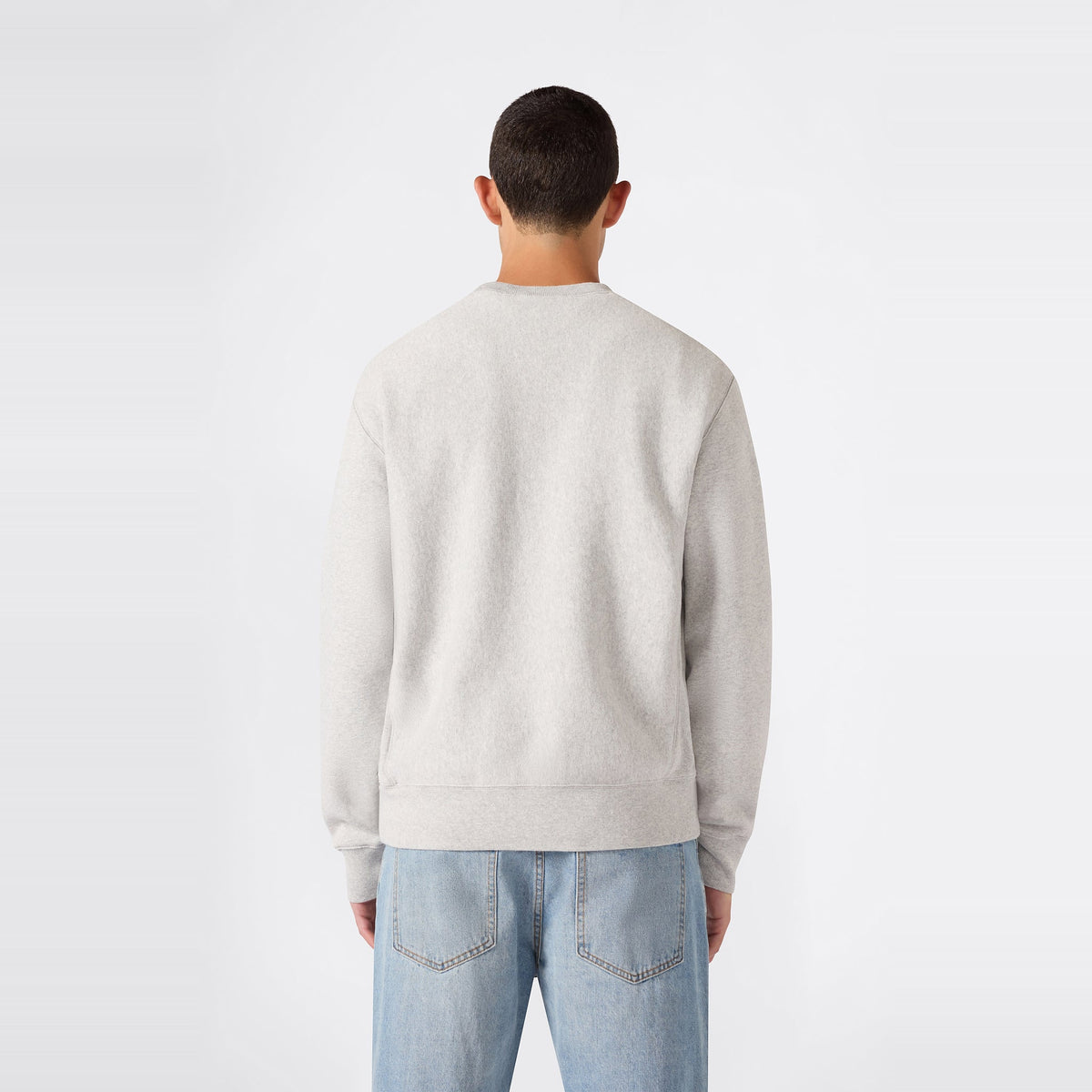 A person with short dark hair is pictured from behind, wearing a light gray Champion Men’s Relaxed Fit Crewneck Sweatshirt and light blue jeans against a plain white background.