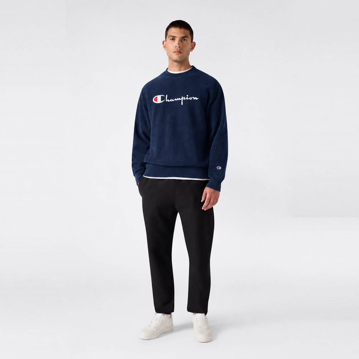 A man wearing a navy blue Champion Men’s Knitted Crewneck Sweatshirt with a vintage embroidered logo layered over a white shirt, paired with black trousers and white sneakers.