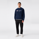 A man wearing a navy blue Champion Men’s Knitted Crewneck Sweatshirt with a vintage embroidered logo layered over a white shirt, paired with black trousers and white sneakers.