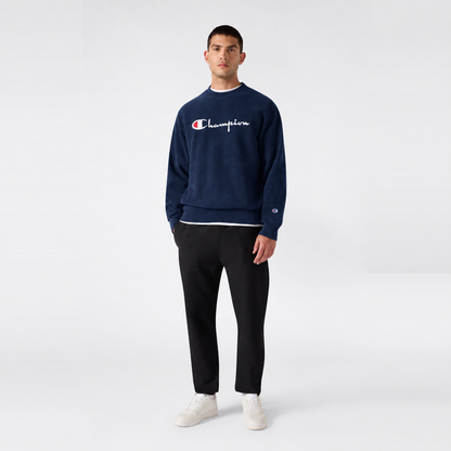 A man wearing a navy blue Champion Men’s Knitted Crewneck Sweatshirt with a vintage embroidered logo layered over a white shirt, paired with black trousers and white sneakers.