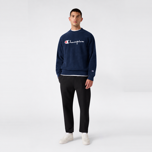 A man wearing a navy blue Champion Men’s Knitted Crewneck Sweatshirt with a vintage embroidered logo layered over a white shirt, paired with black trousers and white sneakers.
