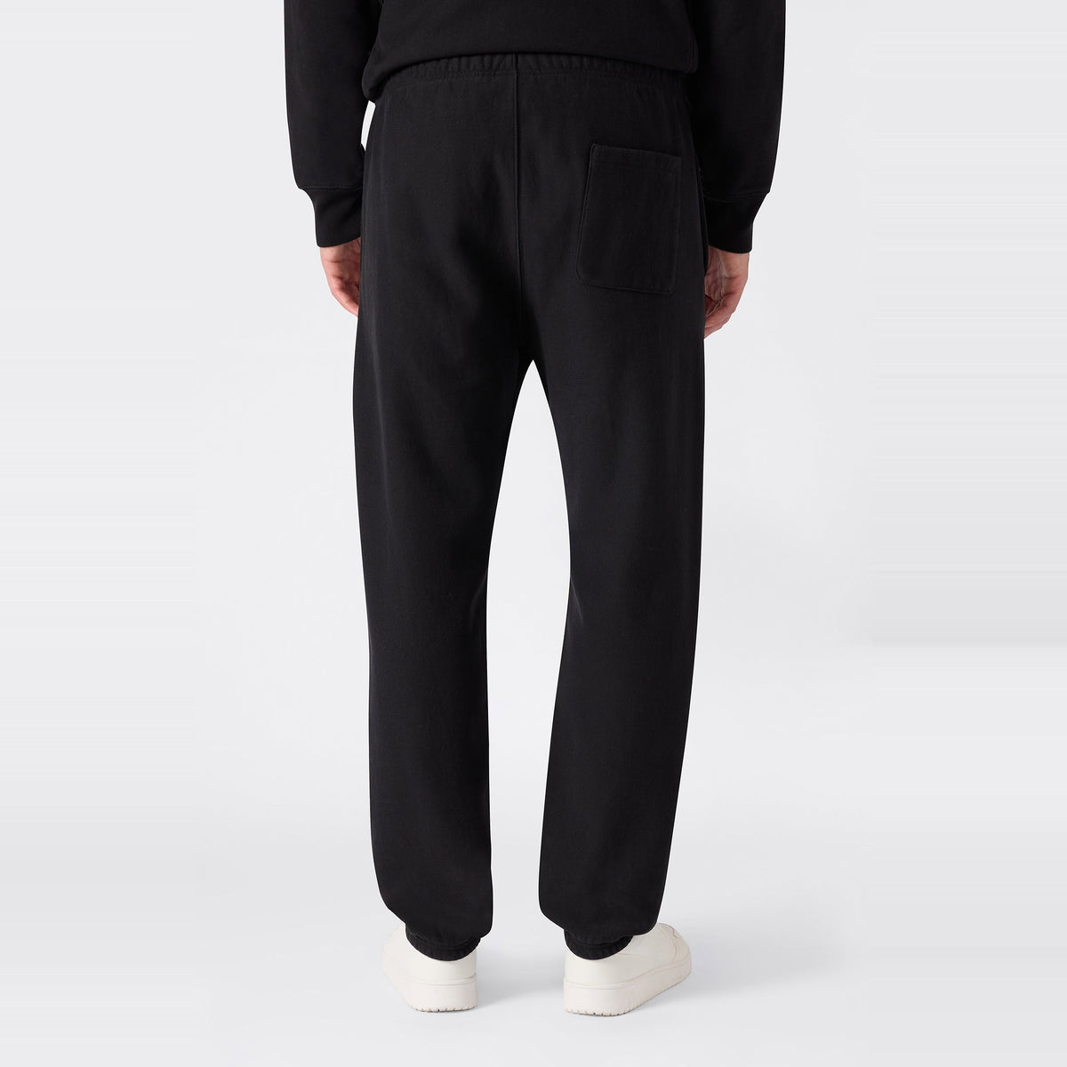 A person is shown from the back against a plain light background, wearing black Champion Men’s Knitted Elastic Cuff Pants—designed in a classic sportswear fleece style with an elastic waistband and single back pocket—and white shoes.