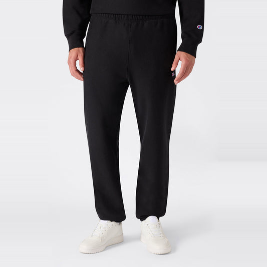 A person is shown from the lower torso down, standing against a plain light background and wearing black Champion Men’s Knitted Elastic Cuff Pants paired with white sneakers. The sportswear fleece sweatpants feature an elastic cuff design and a small Champion logo on the upper thigh.