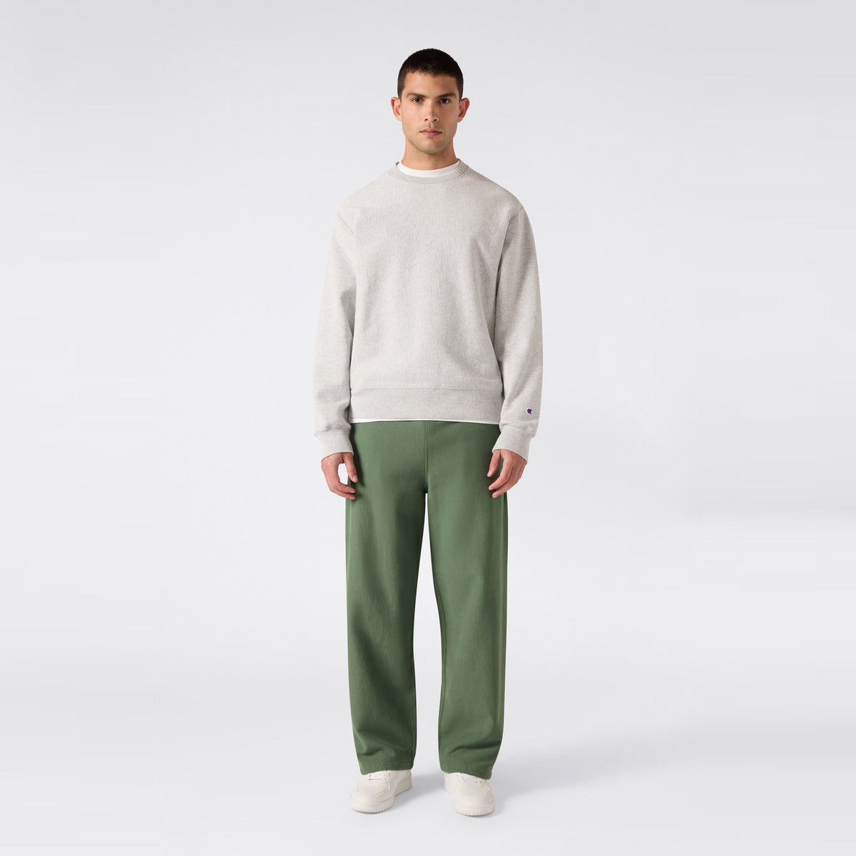 A man stands facing forward against a plain light gray background, wearing a light gray sweatshirt, white shoes, and green Champion Men’s Knitted Wide Leg Pants.