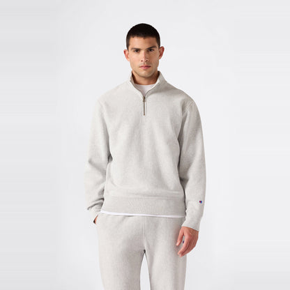 A person stands against a plain background wearing the Champion Men’s Knitted Half Zip Sweatshirt in light gray, styled with matching sweatpants. Crafted from cozy fleece sportswear fabric, the outfit features a relaxed fit and is paired with a casual expression.