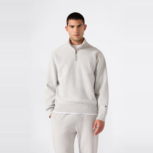A person stands against a plain background wearing the Champion Men’s Knitted Half Zip Sweatshirt in light gray, styled with matching sweatpants. Crafted from cozy fleece sportswear fabric, the outfit features a relaxed fit and is paired with a casual expression.