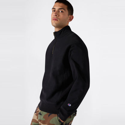 A person wearing a black Champion Men’s Knitted Half Zip Sweatshirt, featuring a small Champion logo on the sleeve and paired with camouflage pants, stands confidently against a plain light background, looking directly at the camera.
