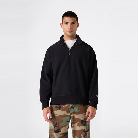 A man faces forward, dressed in a Champion Men’s Knitted Half Zip Sweatshirt in black layered over a white shirt and paired with camouflage pants, set against a plain light background.