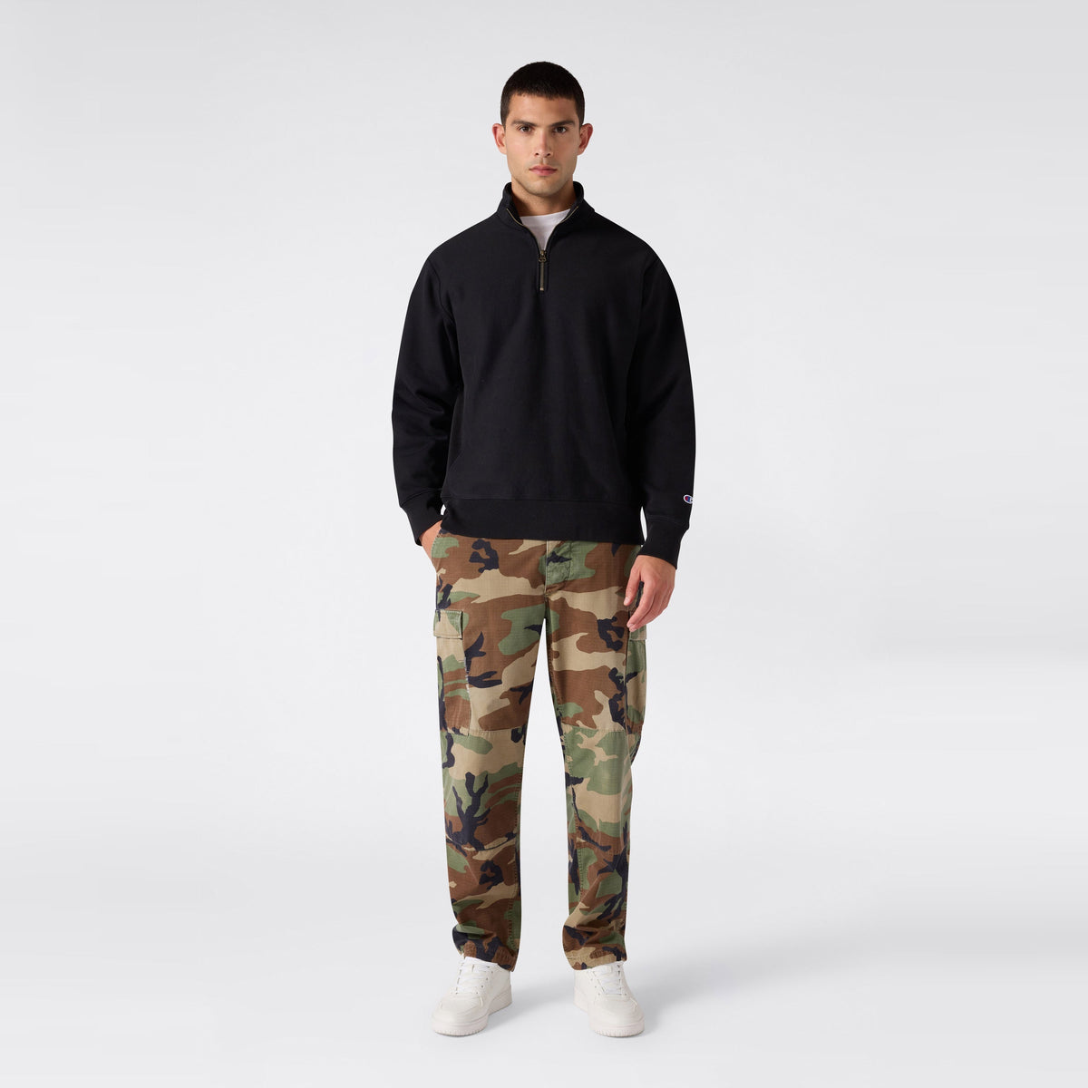 A person stands facing forward wearing a black Champion Men’s Knitted Half Zip Sweatshirt, camouflage cargo pants, and white sneakers against a plain light gray background.