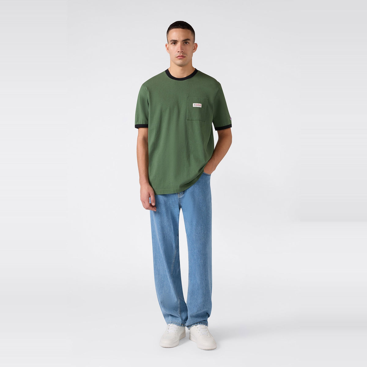 A person stands against a light, plain background wearing the Champion Men’s Knitted Ringer T-Shirt with black trim, paired with light blue jeans and white sneakers. One hand is in their pocket as they look toward the camera.