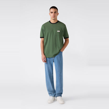 A person stands against a light, plain background wearing the Champion Men’s Knitted Ringer T-Shirt with black trim, paired with light blue jeans and white sneakers. One hand is in their pocket as they look toward the camera.