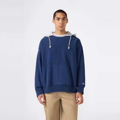 A person stands facing forward against a plain white background, wearing a blue Champion Men’s Knitted Hooded Sweatshirt (Vintage Wash) featuring light gray hood lining and drawstrings, paired with tan pants and a neutral expression.