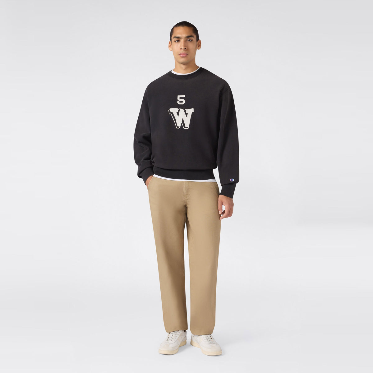 A person stands facing forward against a plain light gray background, wearing a black Champion Men’s Knitted Crewneck Sweatshirt (Reversible + Vintage Wash) featuring “5 W” lettering—crafted from signature Reverse Weave fleece for extra durability—paired with beige pants and white sneakers.