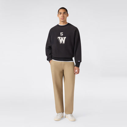 A person stands facing forward against a plain light gray background, wearing a black Champion Men’s Knitted Crewneck Sweatshirt (Reversible + Vintage Wash) featuring “5 W” lettering—crafted from signature Reverse Weave fleece for extra durability—paired with beige pants and white sneakers.