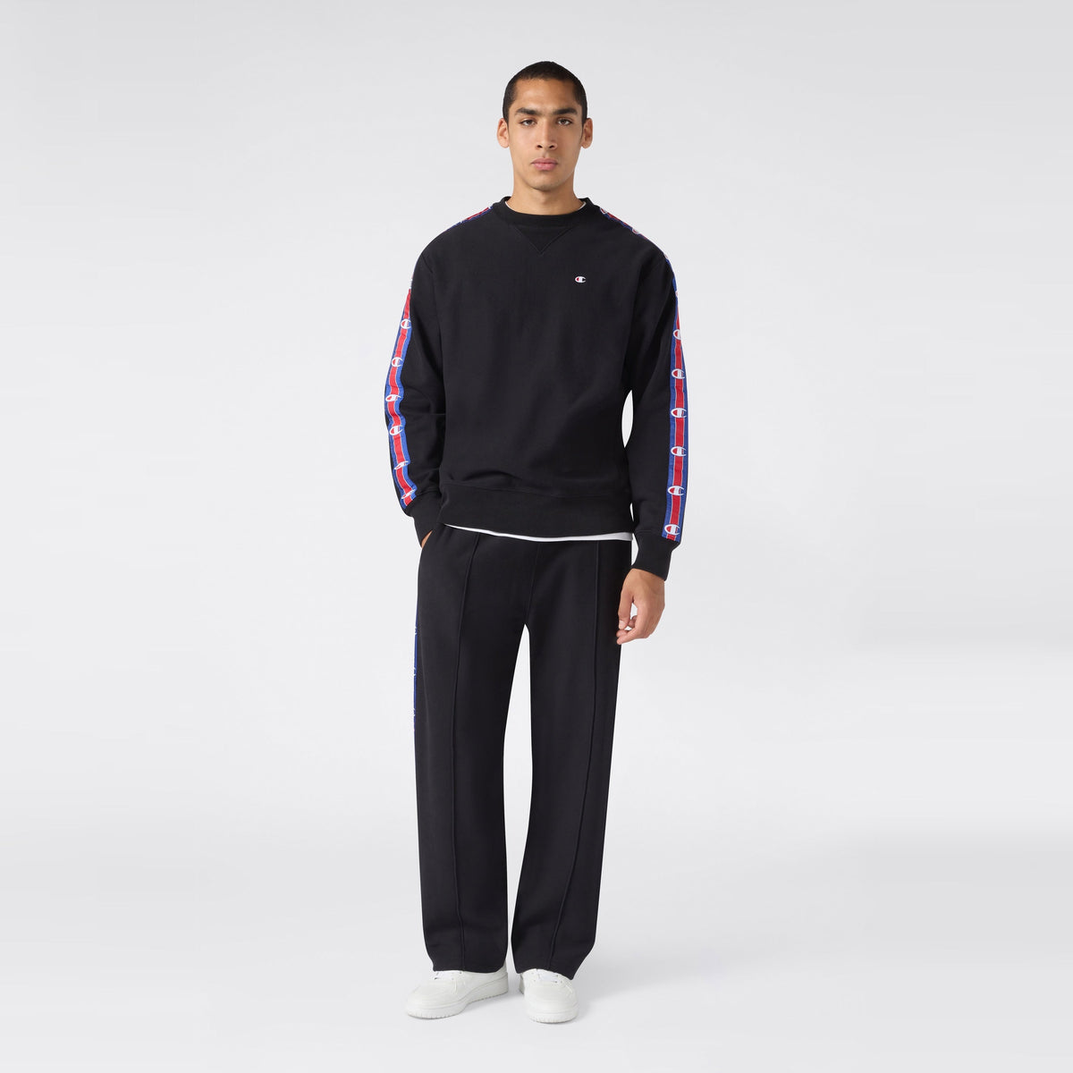 A person stands facing forward, wearing a black sweatshirt with patterned stripes on the sleeves, paired with Champion Men’s Knitted Straight Hem Pants in black featuring matching stripes, and white sneakers, against a plain light background.