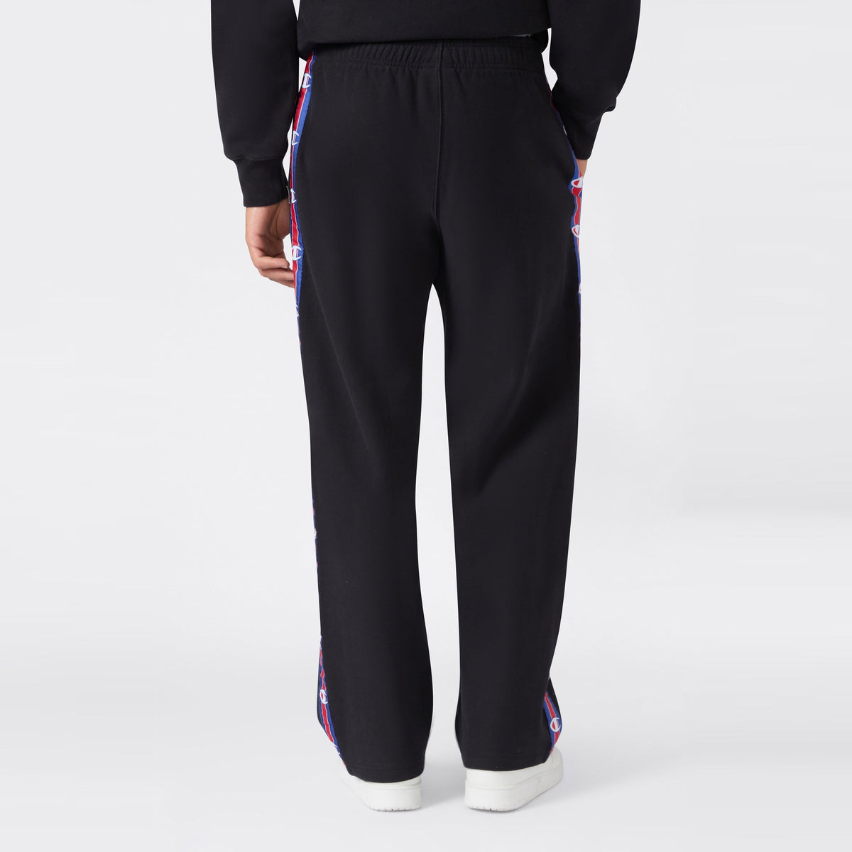 Back view of a person wearing black Champion Men’s Knitted Straight Hem Pants featuring red, white, and blue logo tape along the sides, paired with a matching black sweatshirt and white sneakers against a plain light gray background—an ideal option for men’s sportswear.