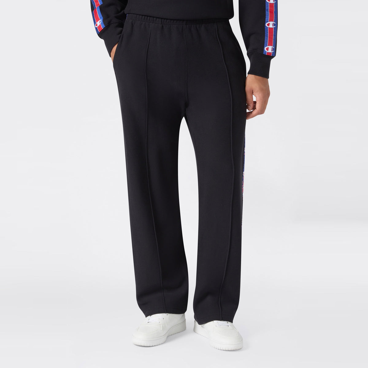 A person wearing black Champion Men’s Knitted Straight Hem Pants paired with white sneakers, standing with hands in pockets against a plain light background—a classic men’s sportswear look.