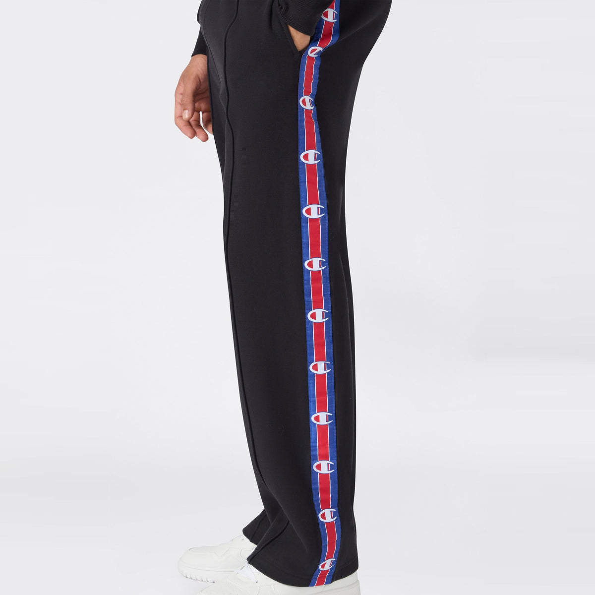 A person wears Champion Men’s Knitted Straight Hem Pants—black trousers with red and blue stripes adorned with Champion logos along the side, styled in classic men’s sportswear fashion—paired with white sneakers. Only the lower body is shown against a plain white background.