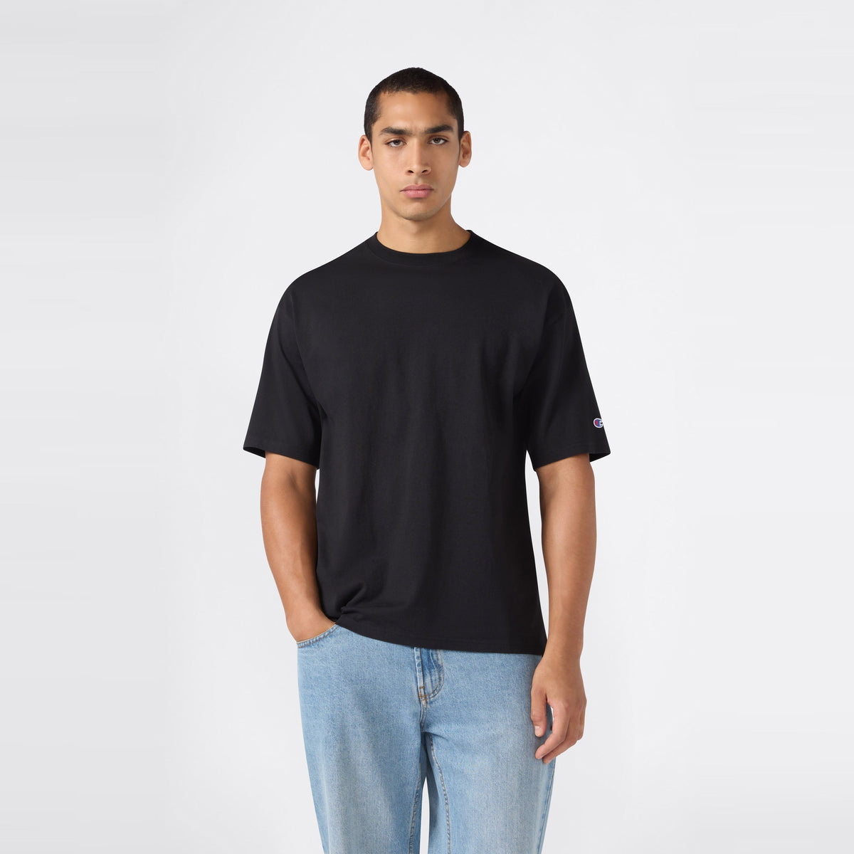 A person wearing a Champion Men’s Knitted T-Shape Tee in plain black and light blue jeans stands against a simple, light gray background, with one hand in their pocket, facing forward.