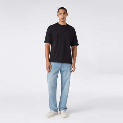 A person stands facing forward, wearing a plain black Champion Men’s Knitted T-Shape Tee, light blue jeans, and white sneakers, against a plain white background.