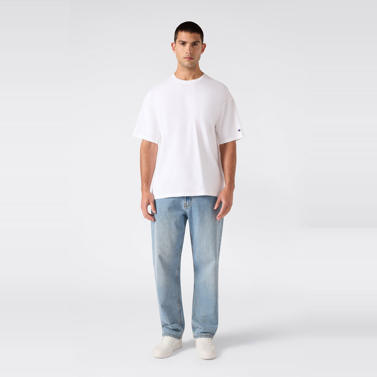 A person stands facing forward, wearing the Champion Men’s Knitted T-Shape Tee, paired with light blue jeans and white sneakers, against a plain light gray background.