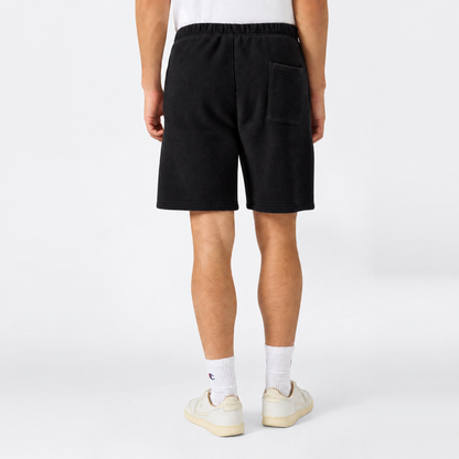 A person stands facing away, wearing a white shirt and black Men’s Knitted Shorts by Champion featuring a back pocket, styled with white socks and off-white sneakers against a plain light background.